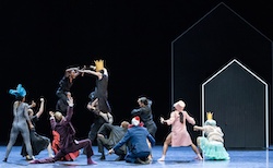 A company of dancers react to a human chicken fight. One woman wears a long-sleeve, form fitting silver lame body suit with a tall, blue feather headdress. Another wears a white, crushed fabric shirt with a light blue tutu and white, fishnet hose. A third wears a long, pink overcoat with a matching, tall, pillbox hat, standing next to a man in white clown makeup with a tiny red top hat and a wide, frilly, red collar and a two-piece navy blue suit.